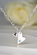 Sterling silver double heart necklace set with aquamarine displayed on white background from the From The Heart collection by Christine Sadler. White rose in the background
