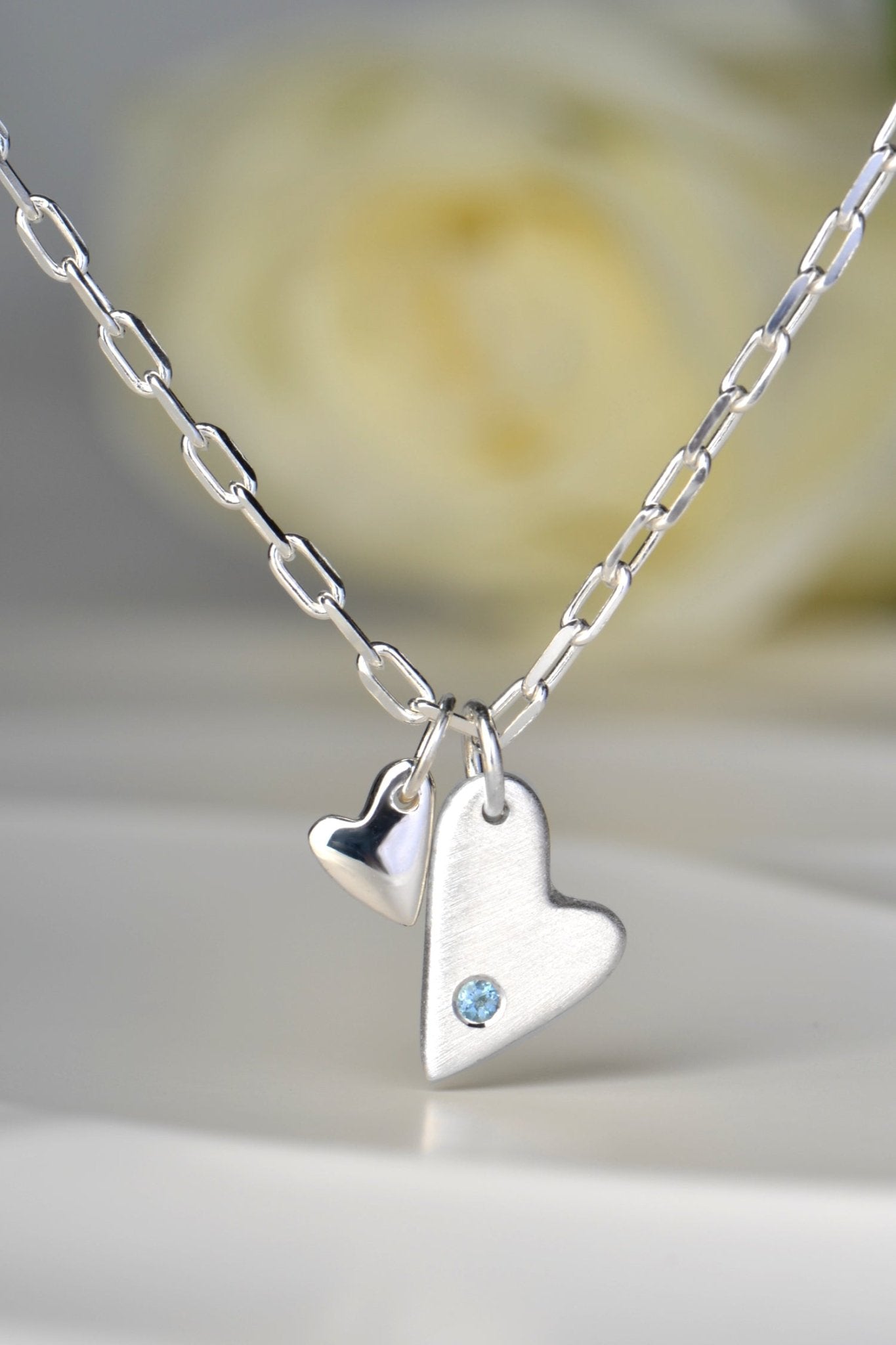 Sterling silver double heart necklace set with aquamarine displayed on white background from the From The Heart collection by Christine Sadler. White rose in the background