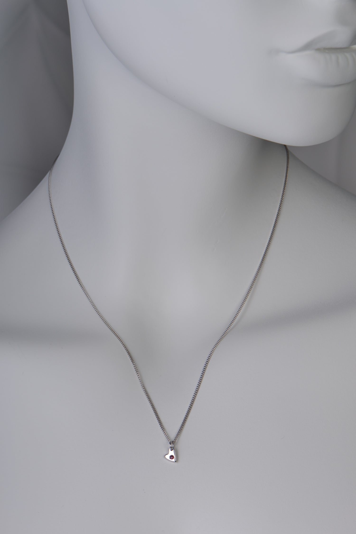 Small polished white gold heart necklace with a ruby displayed on white mannequin to show size and scale when on a 45cm long chain