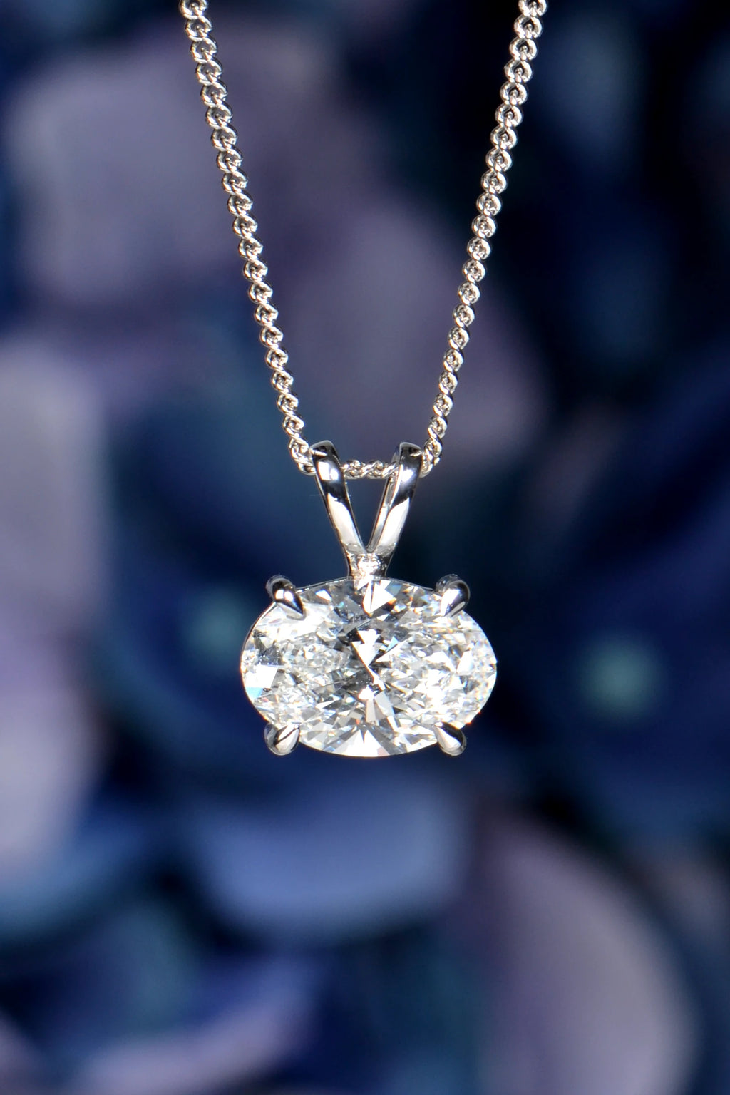 Close-up of a 2.06 carat oval lab-grown diamond pendant necklace in an East-West setting against a blurred blue hydrangea flower background. Christine Sadler
