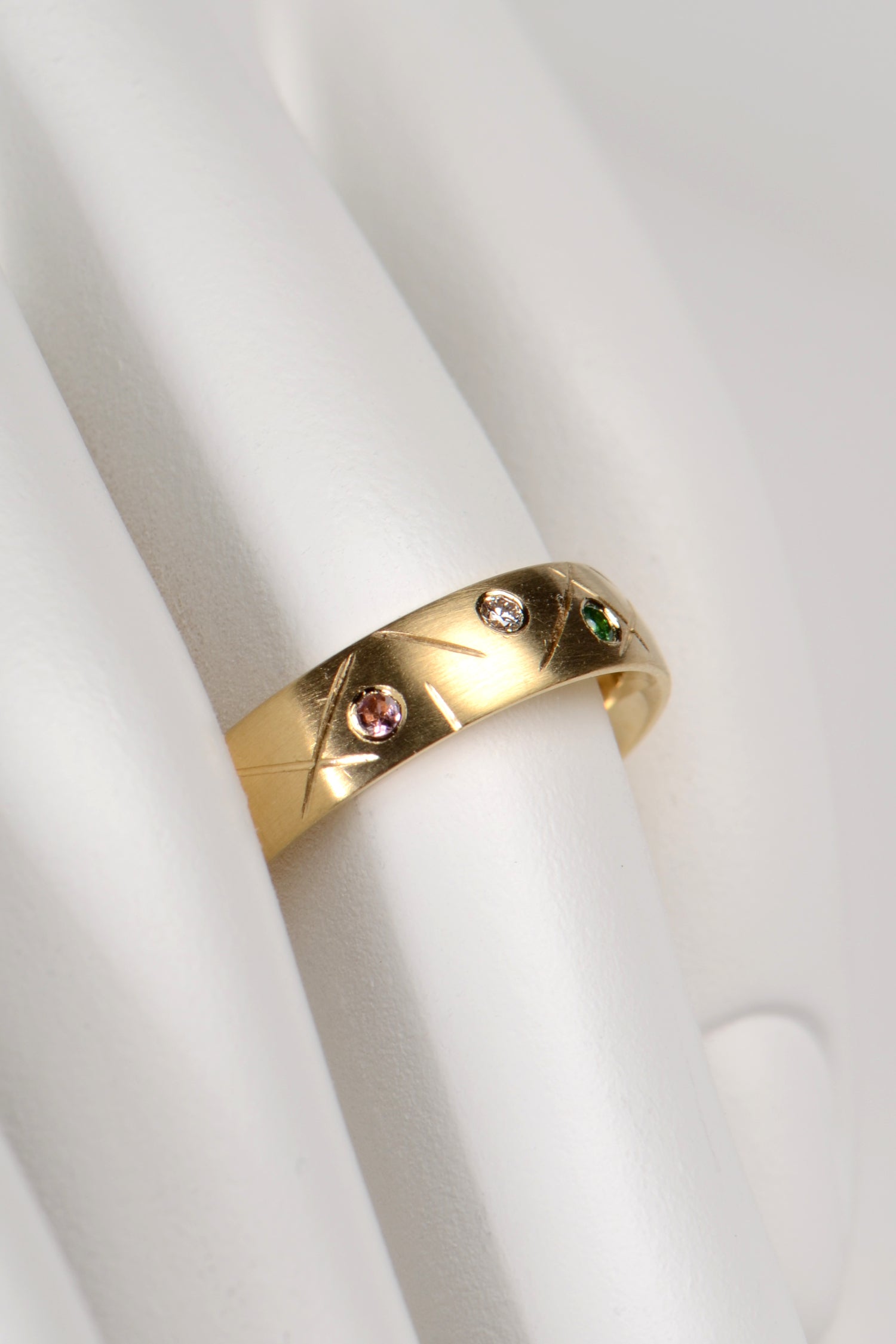 5mm wide 18ct yellow gold ring set with different coloured gemstones all the way round. In this photo we see a pink tourmaline, a green garnet and a diamond. The ring has a criss cross random pattern engraved between the gemstones. By UK jewellery designer Christine Sadler