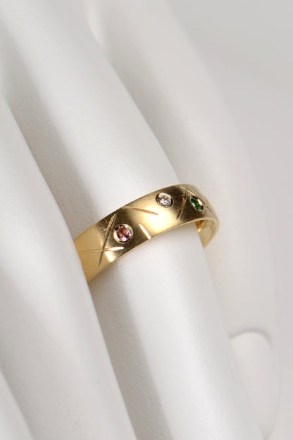 5mm wide 18ct yellow gold ring set with different coloured gemstones all the way round. In this photo we see a pink tourmaline, a green garnet and a diamond. The ring has a criss cross random pattern engraved between the gemstones. By UK jewellery designer Christine Sadler