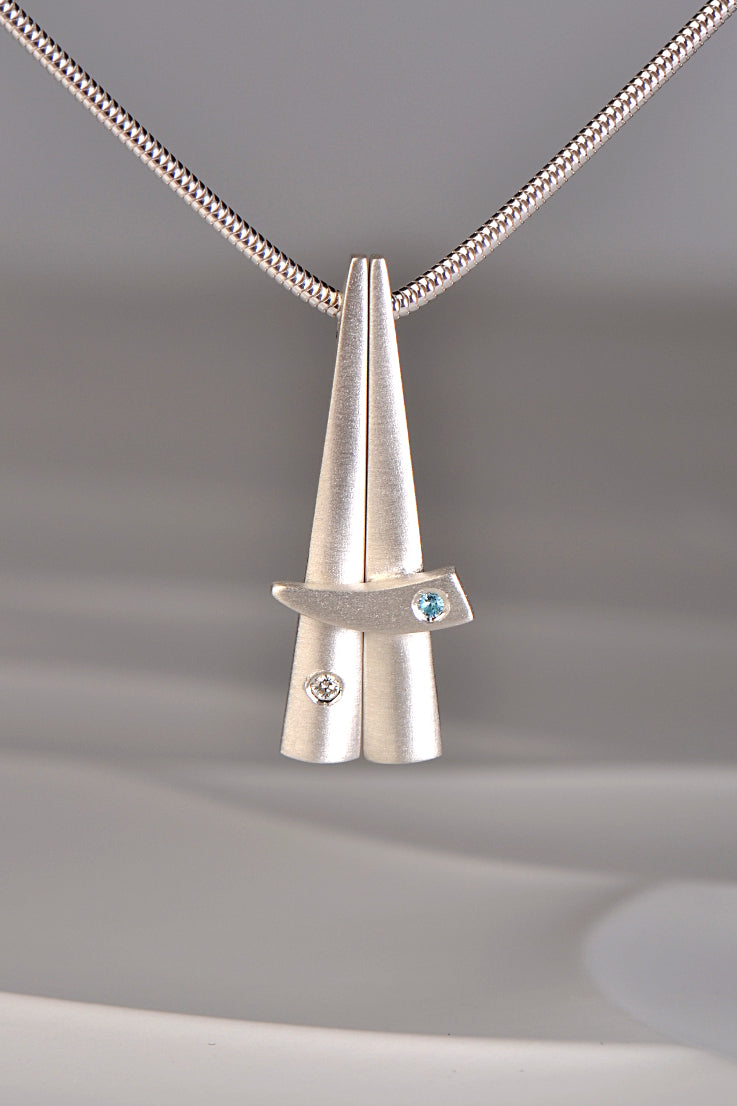contemporary stacking pendants in brushed sterling silver that are inspired by Scottish Cairns by Christine Sadler. One is set with a diamond and the other with a blue topaz.