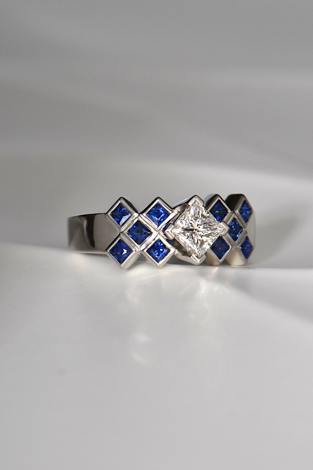 A large central princess cut white diamond is set above a sea of square cut blue sapphires in a platinum engagement ring. There are ten sapphires in total, five on either side of the princess cut central diamond. The Tartan diamond ring is unmistakably Scottish.