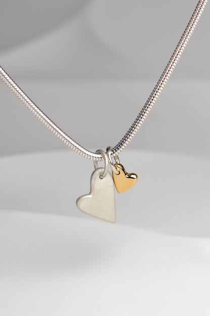 close up of handmade double heart necklace with large matt silver heart and small polished gold heart displayed on white background from the From The Heart collection by Christine Sadler